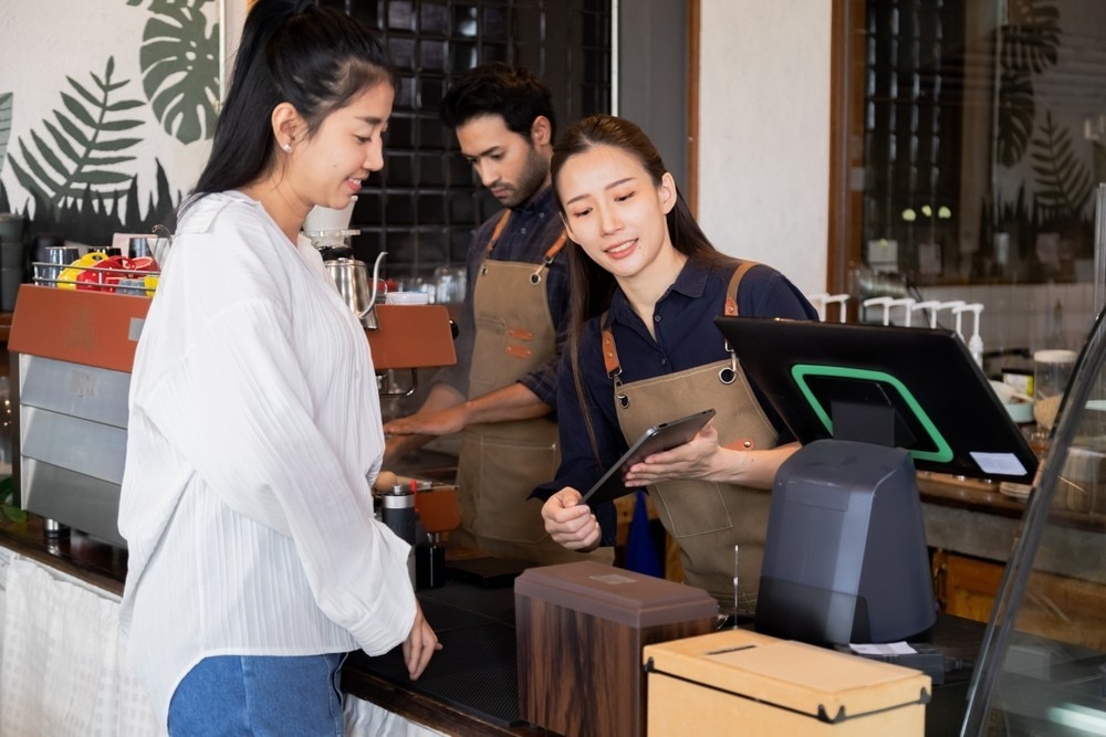 Asian cafe cashier worker female showing menu or cashless billing payment to customer, barista small business owner using technology tablet receives coffee order at workplace in restaurant coffee shop