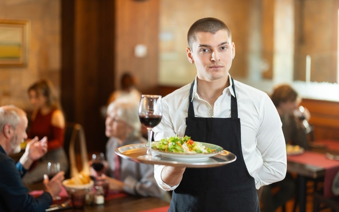 Guy waiter with tray in cafe hall. Employee of public catering cafe carries order to client, ready to serve guest of cafe establishment..