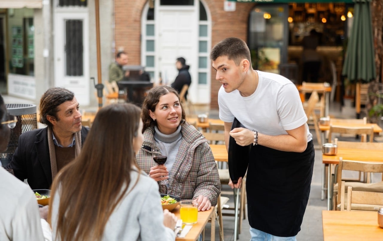 Waiter guy communicates with customers, asks about the quality of food and whether everything is wrong with them. Group of friends ordering food from a waiter in a restaurant