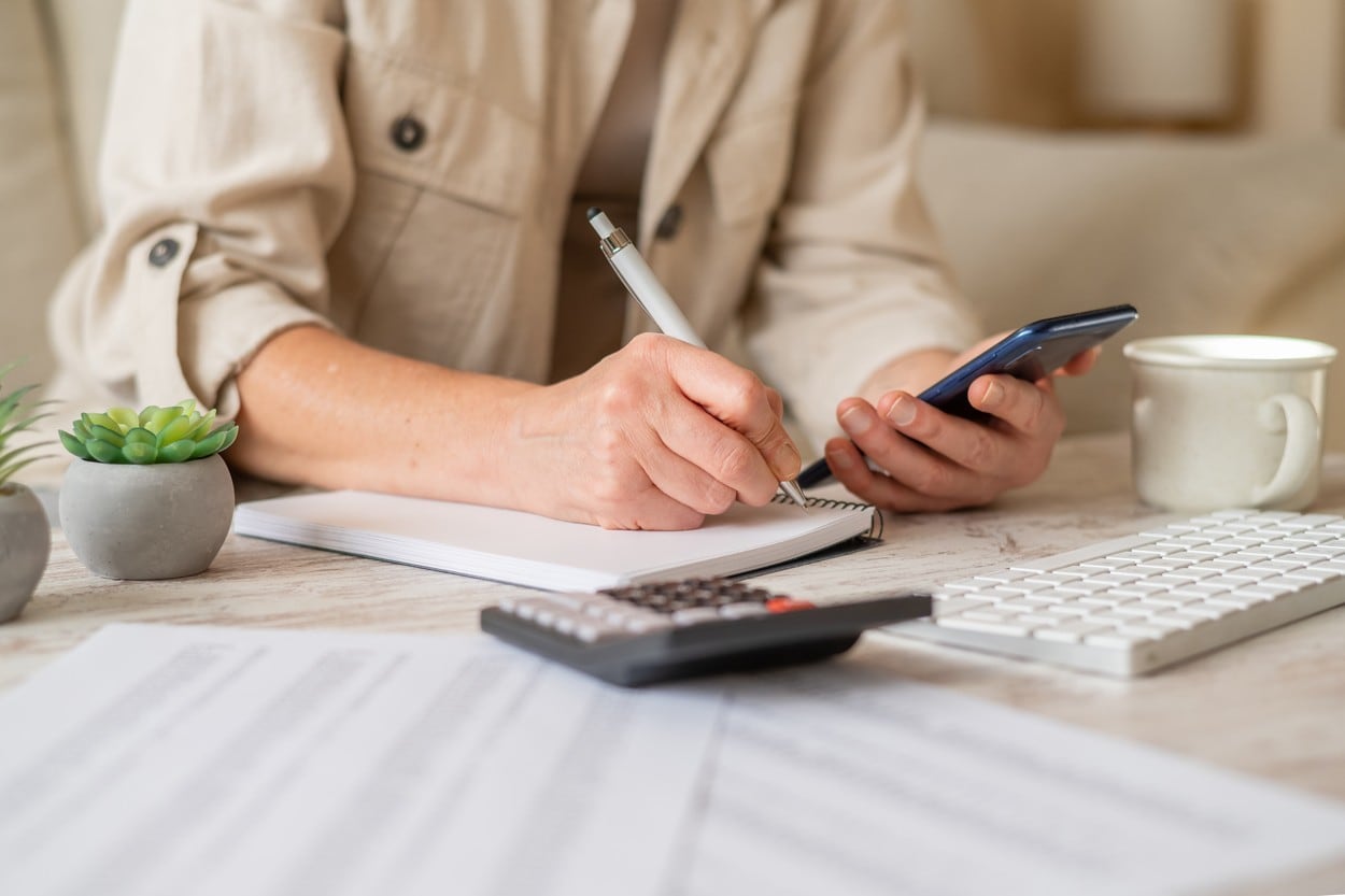 Woman's hands writing notes in a spiral notebook while holding a smartphone, actively managing personal finances, calculating a budget, and planning household expenses on a desk