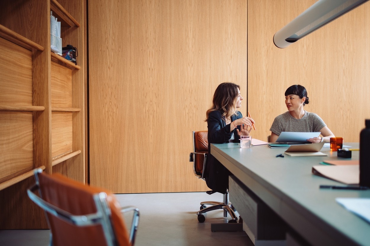 Two businesswomen engaged in a discussion at a modern office table. They are collaborating on a project with documents and laptops.