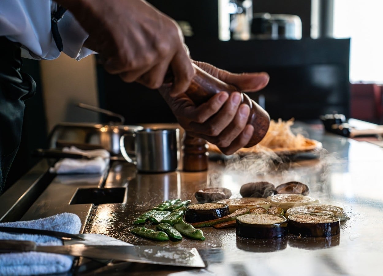 Chef seasoning vegetables on a hot grill in a restaurant kitchen, showcasing culinary skills and fresh ingredients.