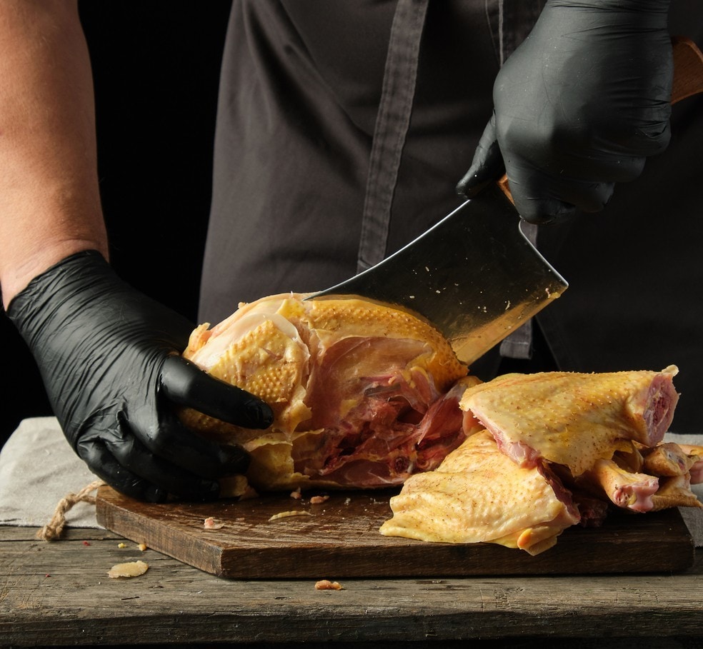 chef in black uniform and latex gloves chopping raw chicken into pieces on a brown wooden board, cooking process