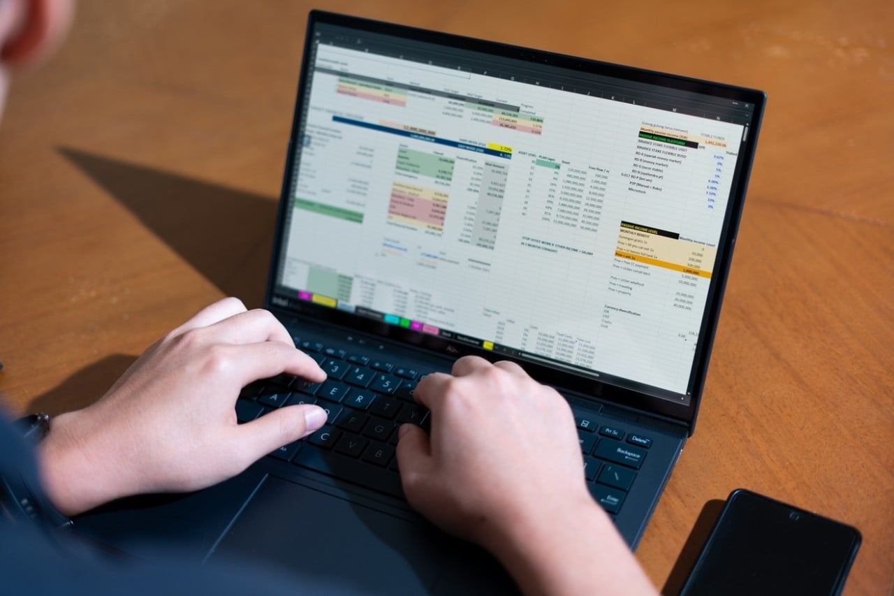 A person is typing on a laptop with a spreadsheet open. The laptop is on a wooden table