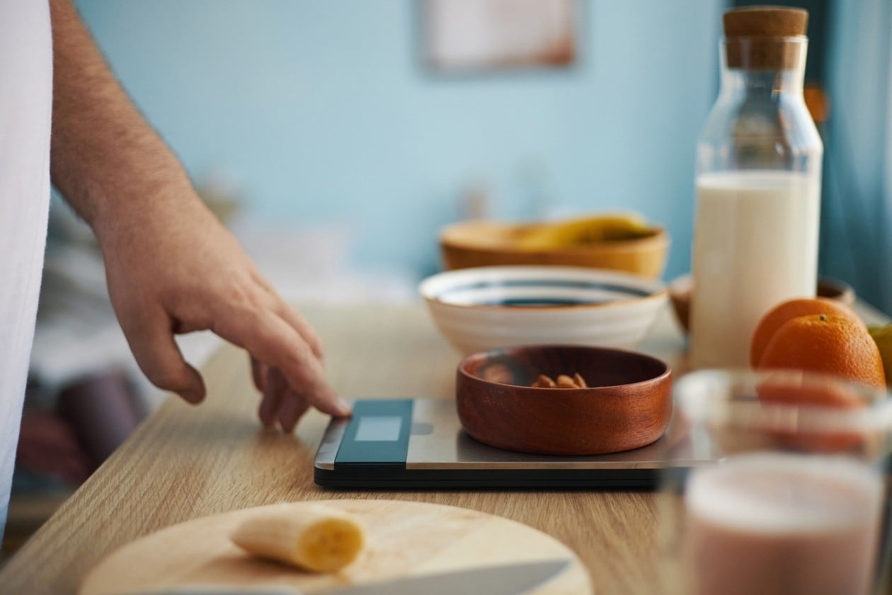 Close-up image showing hand measuring various ingredients on digital kitchen scale, including chopped vegetables, fruits, and liquids in background