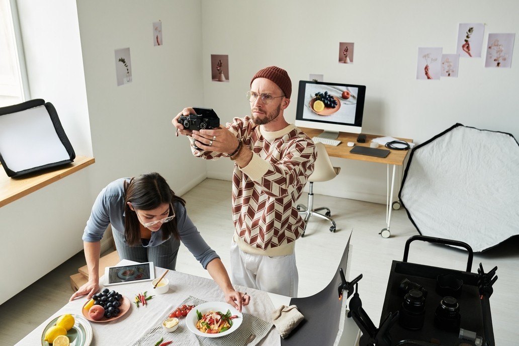 Man holding camera taking photo of food while woman arranging table setting with fruits and salad in bright studio with photography equipment