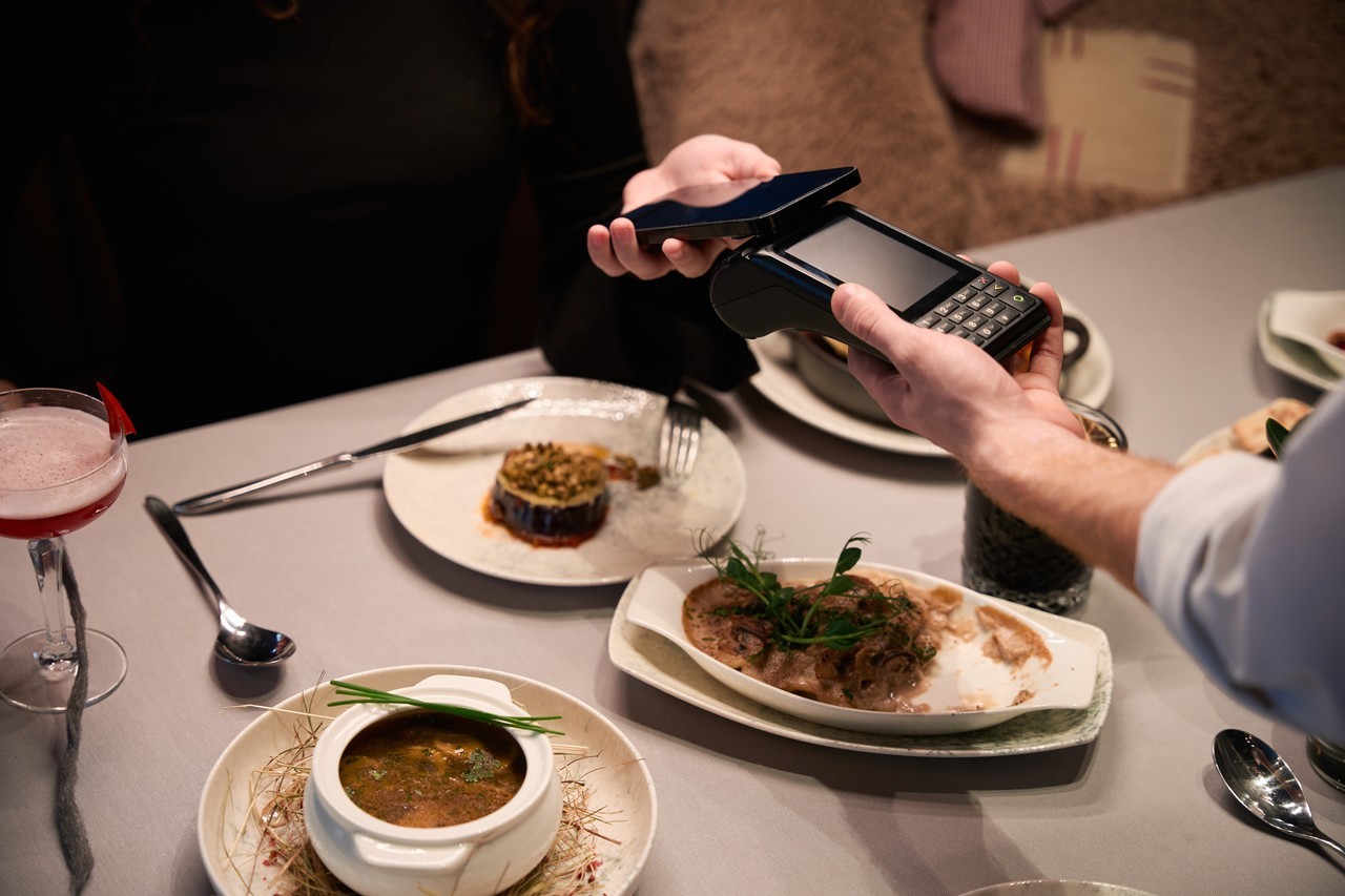 Two people engage in contactless payment at a cozy restaurant in the evening. One person holds a smartphone and payment terminal, while the other assists with the smartphone.