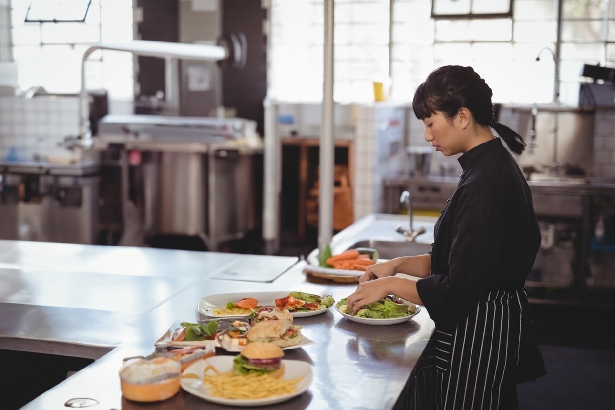 Asian female chef preparing salad in commercial kitchen with plated food and prep table, copy space. Cooking, culinary, restaurant, industrial, stainlesssteel, utensils, natural-light